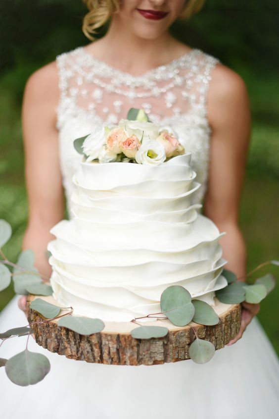 In questa foto una sposa tiene tra le mani una ruffle cake, la torta effetto arricciato. E' poggiata su una base realizzata con il tronco di un albero 