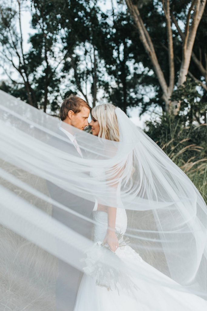 In questa foto due sposi in un bosco si guardano innamorati dietro al velo della sposa tirato in primo piano 