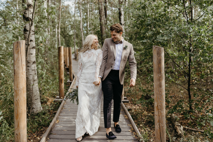 In questa foto due sposi camminano felici mentre si guardano su una pedana di legno e steccato immersa in un bosco. La sposa indossa un abito in pizzo con maniche e tiene un bouquet di felci tra le mani. Lo sposo indossa un abito spezzato marrone e beige e mocassini