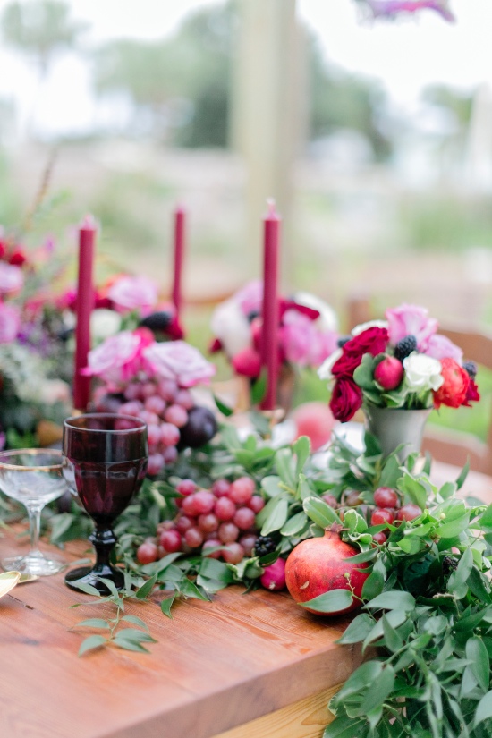 In questa foto il dettaglio di un tavolo matrimonio di legno visto in obliquo perfetto per un matrimonio a settembre. Il runner è realizzato con foglie, uva rossa, melograni e candele bordeaux. Come centrotavola sono usati vasetti di metallo con fiori di campo e ranuncoli rosa, bordeaux e bianchi 