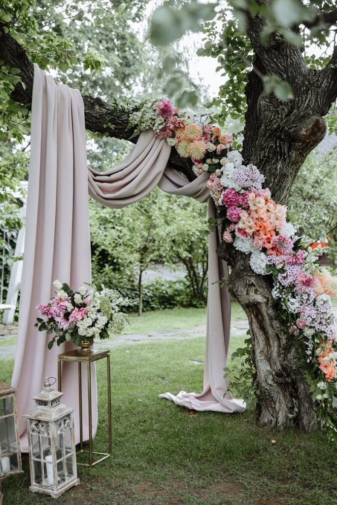 In questa foto un altare per matrimonio civile ricreato sotto un albero di ulivo decorato con drappi di stoffa rosa pallido e fiori fuxia, bianchi rosa pesca. A terra sono disposte alcune lanterne e alzatine dorate