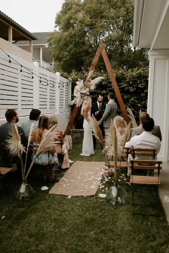 In questa foto un matrimonio civile celebrato su un giardino di casa in stile boho chic. L'allestimento è realizzato con pampas e un triangolo di legno posto dietro all'officiante al posto del tavolo di nozze. Gli sposi si tengono per mano mentre i pochi invitati assistono alla cerimonia