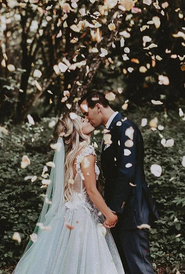 In questa foto due sposi si baciano in un giardino sotto una cascata di petali di rossa rosa. Lei indossa un velo da sposa verde menta