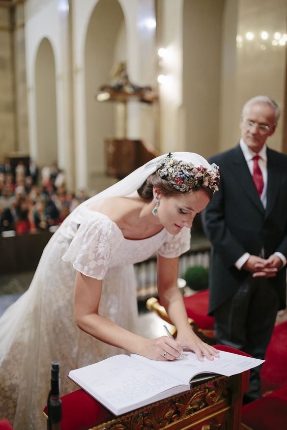 In questa foto una sposa ripresa frontalmente mentre, chinata, firma i documenti del matrimonio in chiesa. In primo piano si vede una folta corona di fiori di campo che circonda l'attaccatura del velo. Indossa una abito con ricami floreali e manichette sulle spalle. La guarda il suo papà dietro di lei con le mani giunte