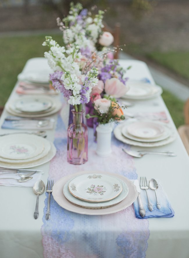 In questa foto una mise en place matrimonio in stile Shabby Chic nei colori del rosa e dell'azzurro. Il tavolo rettangolare è ripreso frontalmente ed è decorato con un runner dipinto in watercolor e tovaglioli ricamati abbinati. Al centro del tavolo sono disposti vasetti e bottigliette rosa, viola e bianchi con fiori di campo. La mise en place è completata con porcellane e posate in argento vintage