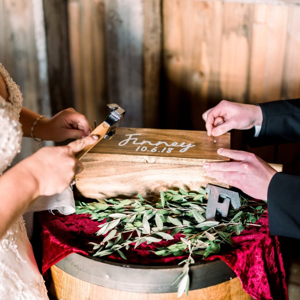 In questa foto due sposi chiudono una scatola di legno durante il rito simbolico per matrimonio della scatola della memoria. La scatola è poggiata su una botte di legno decorata con un drappo di velluto rosso e rami di ulivo. Sulla scatola è riportata la data delle nozze, 10-06-2018. Lo sposo tiene i chiodi mentre la sposa li pianta con un martello