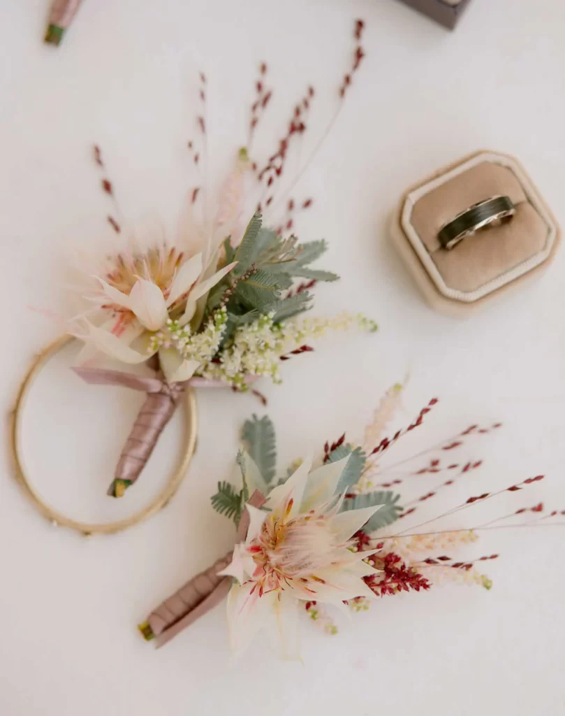 In questa foto due bottoniere con fiori matrimonio di fiori bianchi a forma di stella con felce posizionati accanto alla fede dello sposo