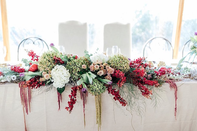 In questa foto un runner floreale su un tavolo per matrimonio rettangolare composto da fiori di amaranto rossi, rose cipria, ortensie bianche, melograni