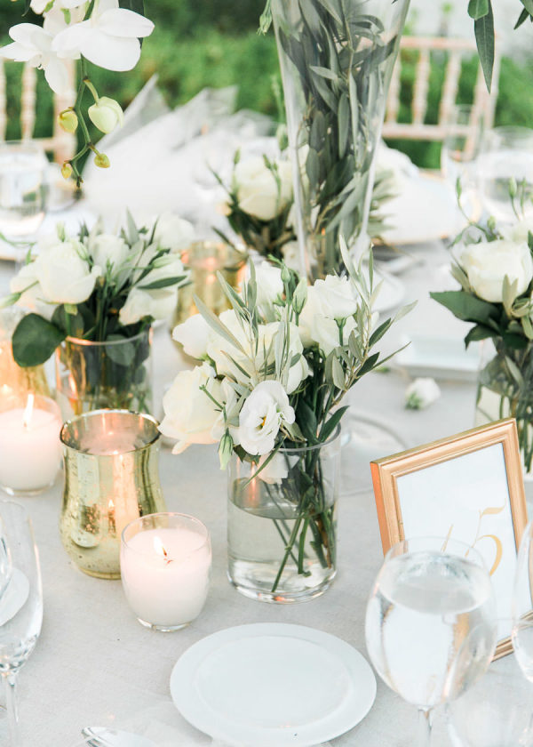 In questa foto un tavolo matrimonio allestito con vasetti pieni di fiori di lisianthus bianchi e foglie di ulivo. Sul tavolo sono presenti anche portacandele bianche e colore oro e una cornice colore oro con il numero del tavolo
