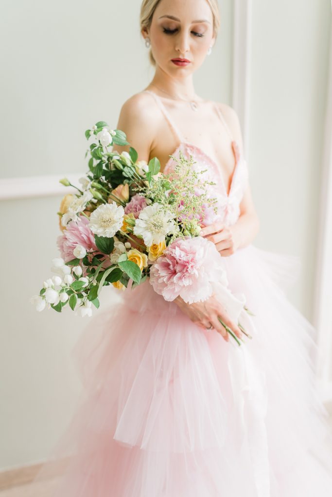In questa foto una modella con un vestito di tulle rosa tiene sull'avambraccio destro un bouquet da sposa con fiori grandi rosa, bianchi e gialli