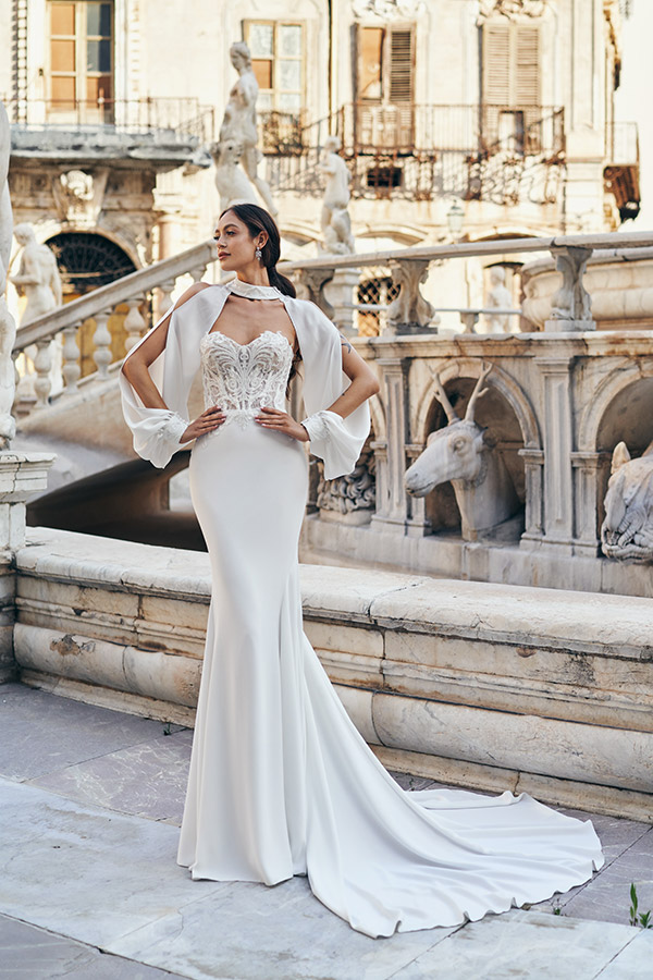 In questa foto una modella indossa un abito da sposa a semi sirena di Annais Bridal in crêpe georgette, bustier di pizzo con scollo a cuore e collare gioiello con maniche rimovibili e semi aperte