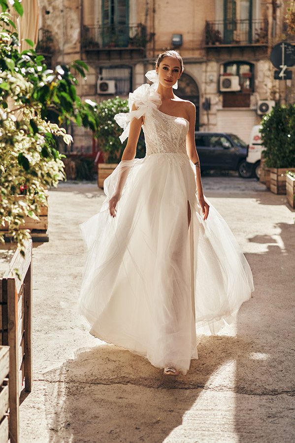 In questa foto una modella cammina per strada indossando uno degli abiti da sposa Annais Bridal 2023 in tulle con corpetto monospalla con maxi fiore 3D sulla spalla destra