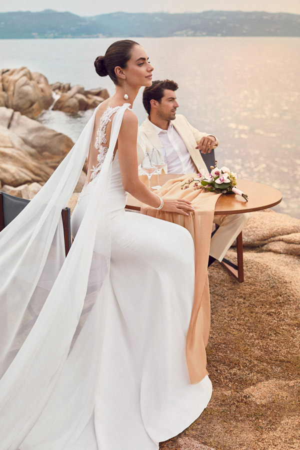 In questa foto due sposi seduti ad un tavolo su scoglio sul mare. In primo piano è presente la sposa che indossa un abito semi sirena in crêpe con scollatura illusion sulla scuola e drappi che scendono dalle spalle