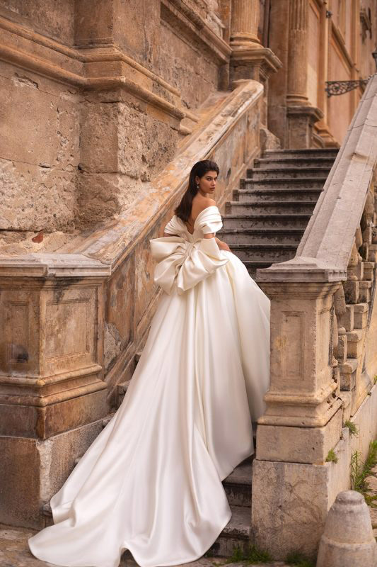 In questa foto una modella, ritratta mentre sale le scale, indossa un abito da sposa con maxi fiocco sulla schiena della linea Eva Lendel 2023