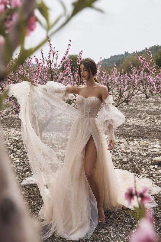 In questa foto una modella in un campo di ciliegi mentre indossa un abito da sposa con spacco e maniche balloon rimovibili di Lee Grebenau 2023