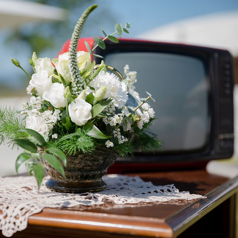 In questa foto un vecchio televisore in bianco e nero come arredo riciclato nozze green. Davanti all'apparecchio una composizione floreale sui toni del bianco e verde, in un vaso bronzato stile vintage