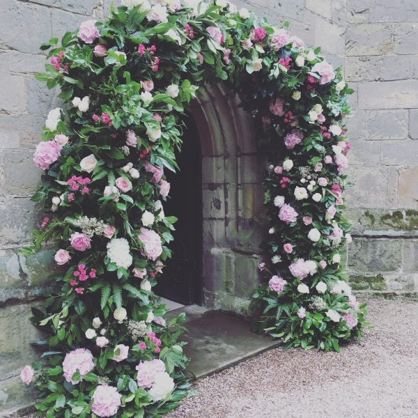 In questa foto, un arco floreale di ortensie, rose inglesi, roselline e foglie verdi decora l'ingresso di una chiesa 