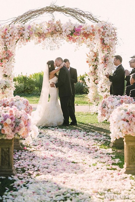 In questa foto, una coppia di sposi si bacia davanti un arco di fiori e banchi all'aperto decorati con composizioni di fiori in bianco e rosa brillante