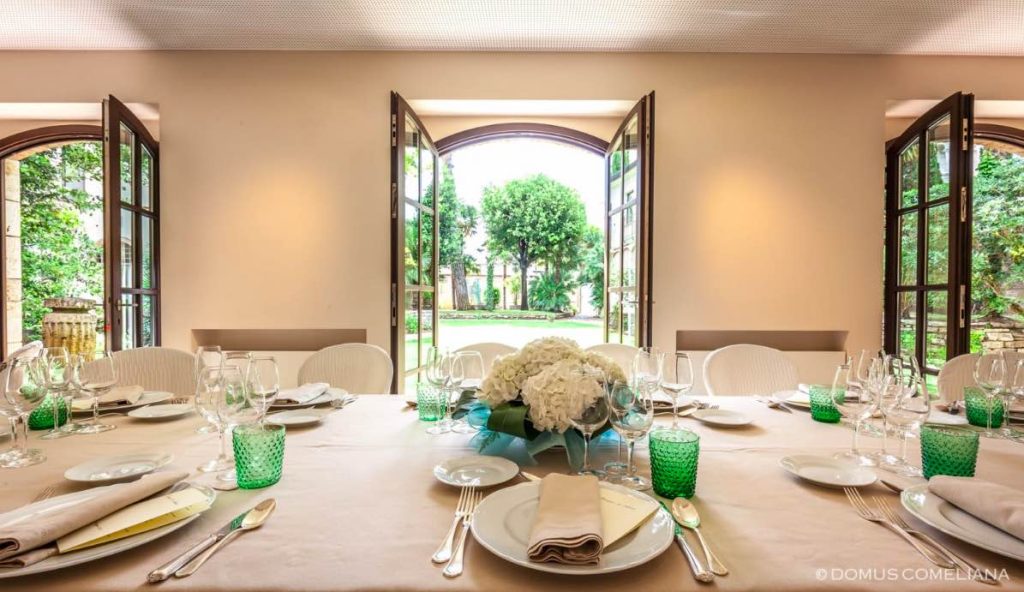 In questa foto un tavolo apparecchiato per un pranzo di nozze, nei toni del verde, posizionato davanti alle grandi porte-finestre aperte che danno sul giardino della Domus Comeliana