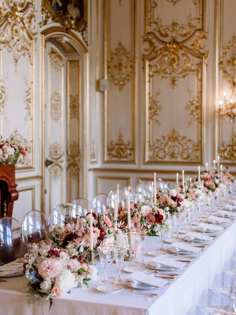 In questa foto un tavolo imperiale in una grande sala di una dimora storica con pareti di colore bianco e oro. Il tavolo è decorato con fiori di colore rosa e rosso e candele