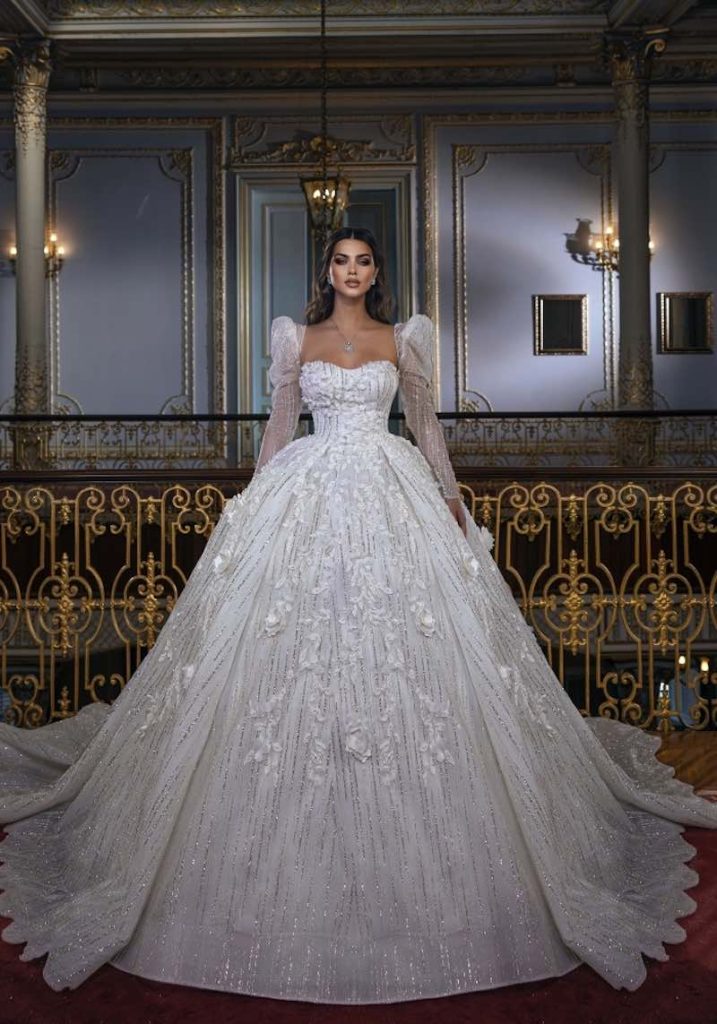 In questa foto una modella indossa un abito da sposa Dovita Bridal 2023 dal volume maxi ricamato con glitter e fiori 3D. L'abito presenta una scollatura a cuore e maniche lunghe gigot