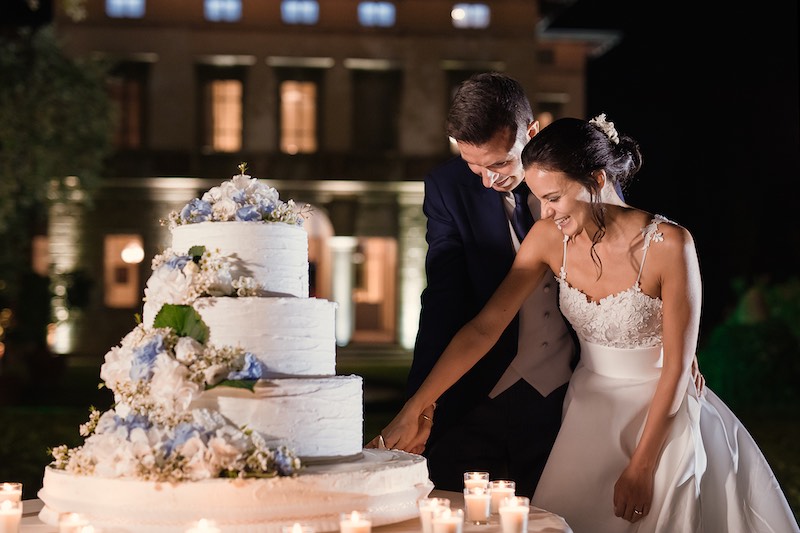 In questa foto di Nicola Tanzella due sposi tagliano una torta nuziale a tre piani decorata con fiori di colore bianco e azzurro. Il tavolo è decorato con piccole candele 