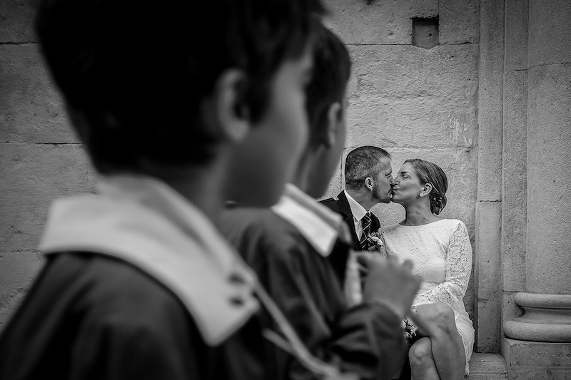 In questa foto in bianco e nero di Nicola Tanzella in primo piano due bambini fuori fuoco e due sposi si baciano seduti su un gradino si baciano