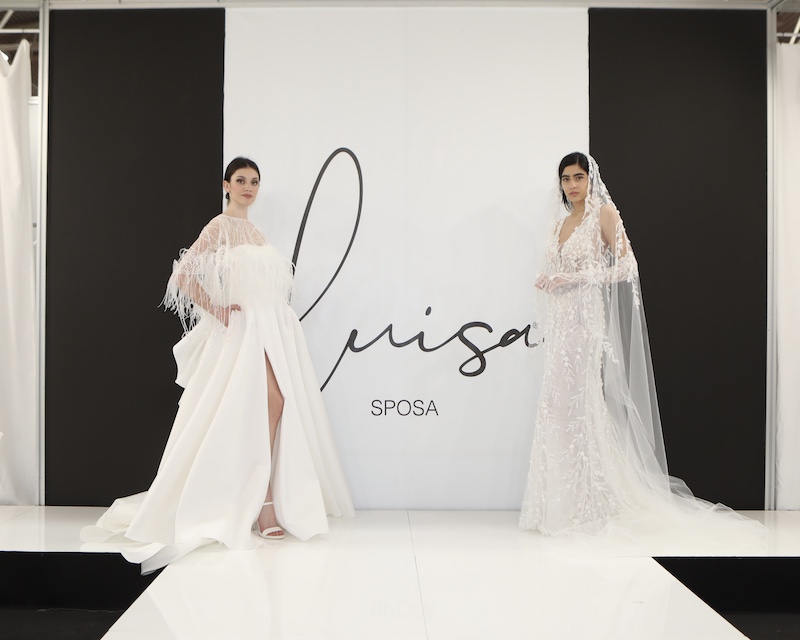 In questa foto due modelle indossano due abiti da sposa Luisa Sposa 2024. A sinistra una modella indossa un abito ampio in mikado con spacco e cappa plumage. A destra una modella indossa un abito a sirena con pizzo floreale e velo abbinato