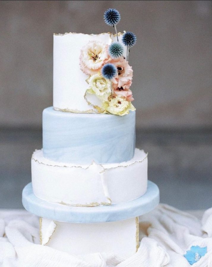 In questa foto una torta nuziale a 3 piani di colore bianco e con effetto watercolor di colore azzurro polvere al centro. La decorazione è completata da fiori di colore rosa, blu e giallo