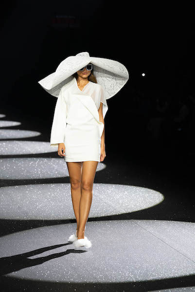 In questa foto una modella sfila indossando un abito bianco corto con cappello a maxi falda della collezione Carlo Pignatelli 2024