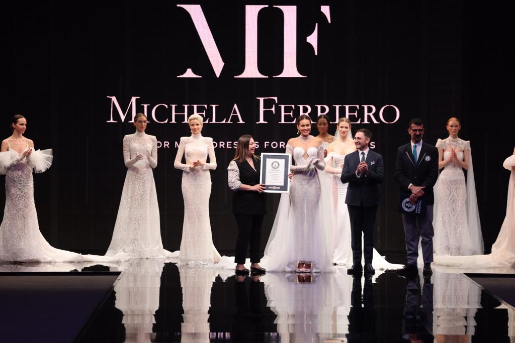 In questa foto la designer Michela Ferriero e il Ceo dell'azienda, Lorenzo Albanese, ricevono l'attestato del Guinness World Record sulla passerella del Sì Sposaitalia circondati dalle modelle che indossano gli abiti da sposa Michela Ferriero 2024