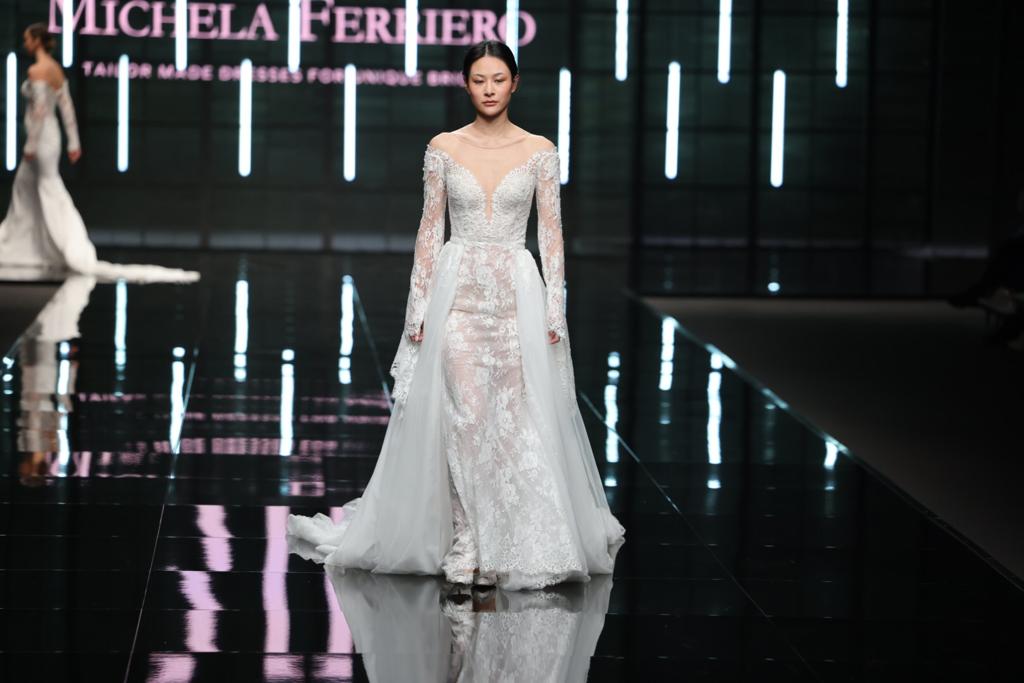 In questa foto una modella sfila indossando un abito da sposa Michela Ferriero 2024 a sirena in pizzo con maniche lunghe, scollo a cuore e sopra gonna in tulle