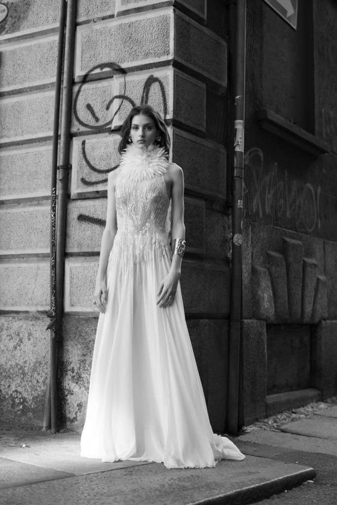 In questa foto in bianco e nero una modella indossa un abito da sposa con gonna in chiffon e corpetto ricamato con perline e collo alto plumage de Le Fate Milano