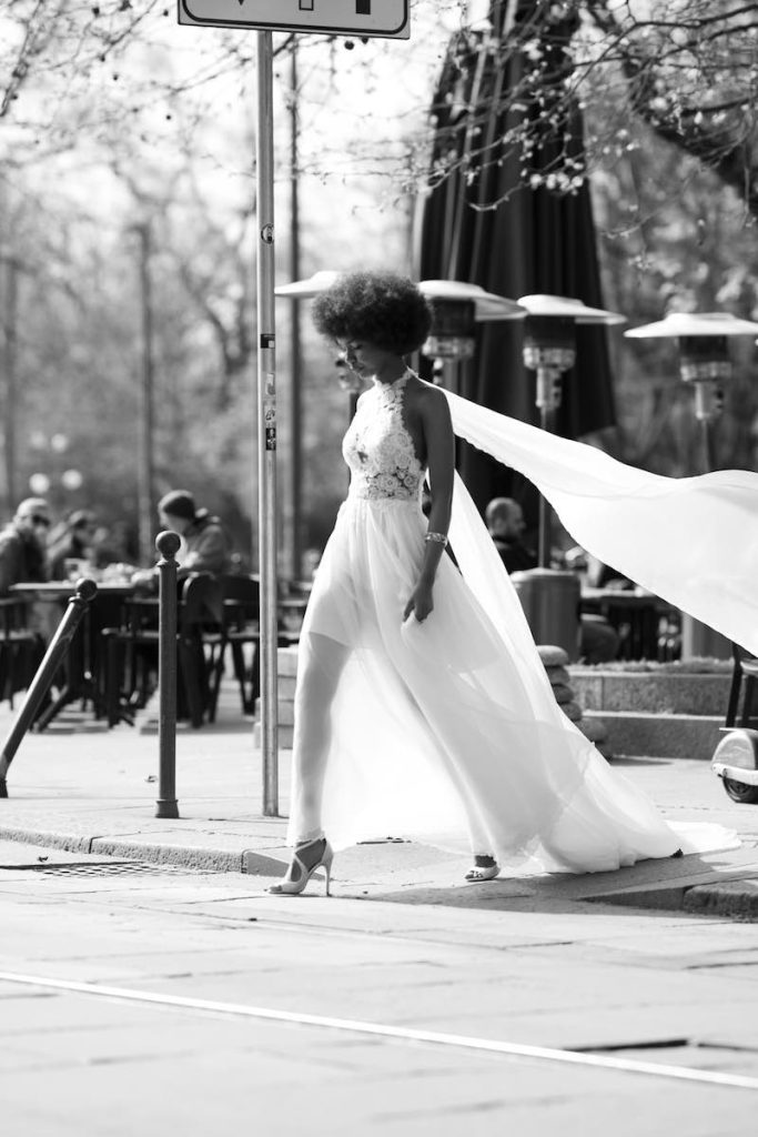 In questa foto in bianco e nero una modella cammina per le strade di Milano indossando un abito da sposa in chiffon creponne con corpetto in pizzo e lunghe ali fluttuanti Le Fate Milano 2024