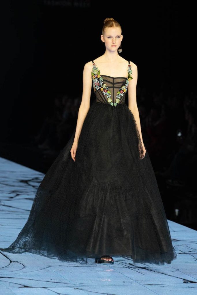 In questa foto una modella sfila indossa un abito da sposa Marco y Maria 2024 di colore nero con gonna ampia e ricami di fiori e trasparenze sul busto 