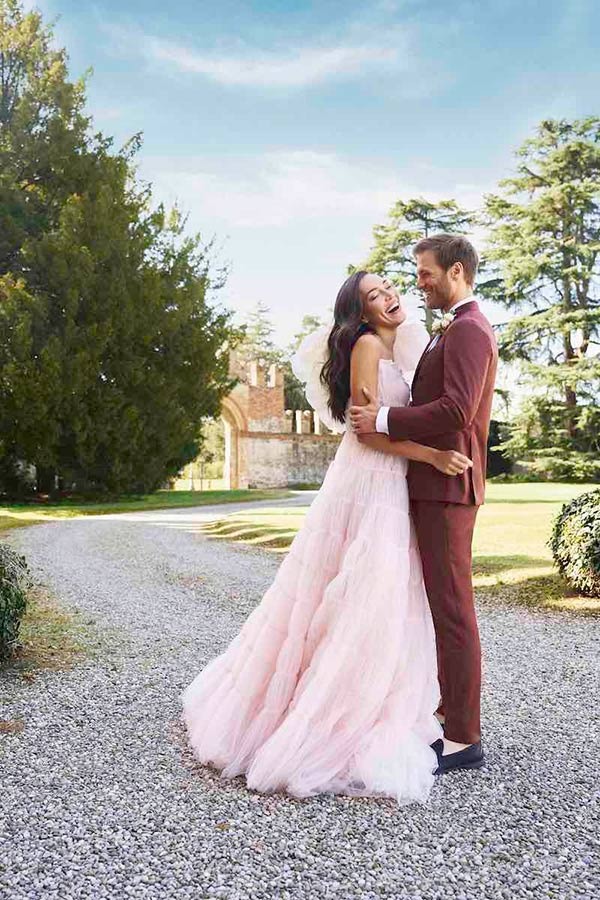 In questa foto due sposi si abbracciano felici nel parco di un'antica dimora. Lui indossa un abito colo borgogna. Lei un abito da sposa di colore rosa con un maxi fiore sulla spalla sinistra