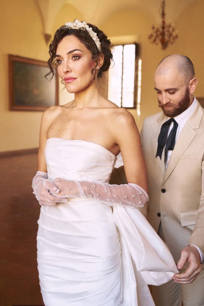 In questa foto il Wedding Planner Maurizio Lo Greco sistema l'abito della sposa che indossa un abito corto drappeggiato e un paio di guanti di tulle con perline