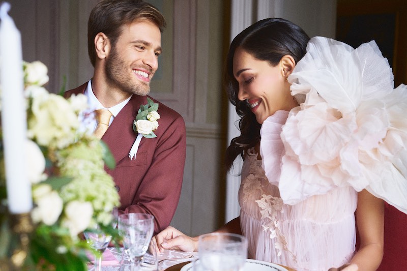 In questa foto due sposi seduti al tavolo di nozze si sorridono. Lui indossa un abito colo borgogna. Lei un abito da sposa di colore rosa con un maxi fiore sulla spalla sinistra