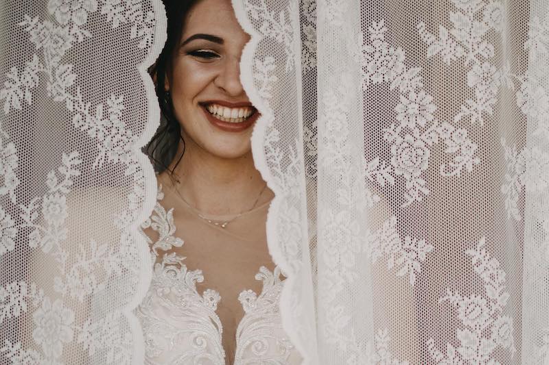 In questa foto di Alberto Cosenza il viso di una sposa che sorride inquadrata attraverso una tenda ricamata in pizzo