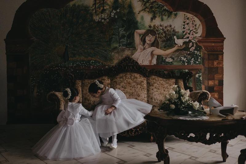 In questa foto di Alberto Cosenza due piccole damigelle in abito di tulle e con uno chignon decorato da nebbiolina bianca giocano su un divano antico in attesa della sposa. In primo piano si intravede il bouquet e sullo sfondo un dipinto sulla parete 