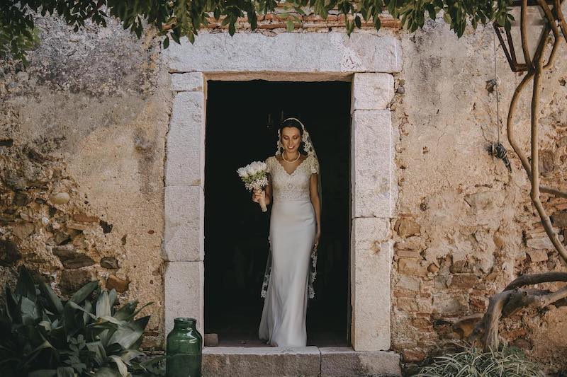 In questa foto di Alberto Cosenza Photographer una sposo con abito a sirena ricamato, velo a mantiglia e un bouquet di fiori bianchi mentre attraversa la porta di una casa in pietra