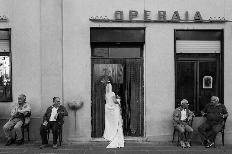 In questa foto in bianco e nero una sposa mentre entra in un negozio con l'insegna "Operaia" mentre fuori, su delle sedie, sono seduti quattro uomini che chiacchierano