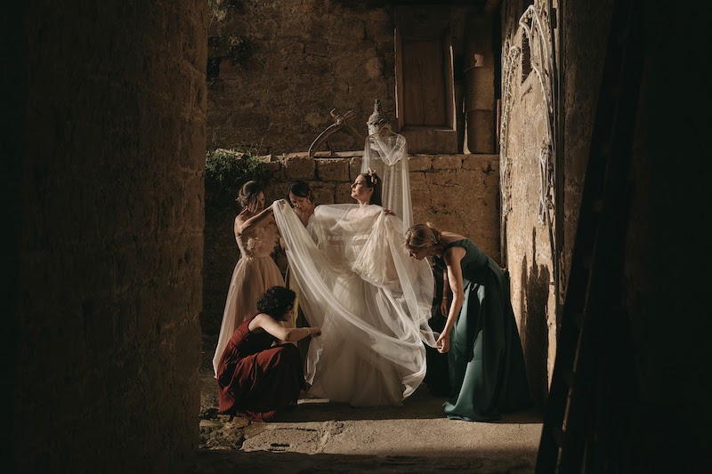 In questa foto di Alberto Cosenza Photographer una sposa tra le vie di un paese durante la vestizione aiutata da quattro damigelle