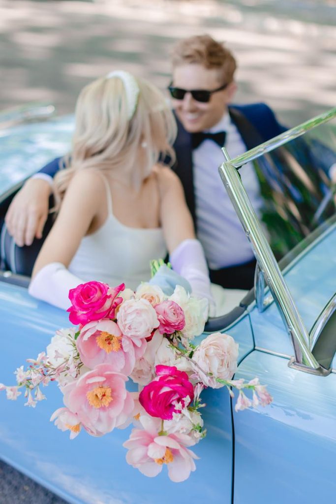 In questa foto due sposi su un'auto decappottabile di colore azzurro si guardano e ridono mentre la sposa tiene fuori dal finestrino un bouquet di fiori di colore rosa, fucsia e bianco