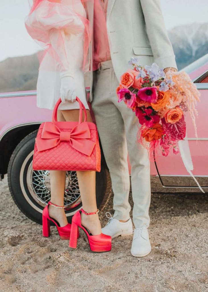 In questa foto due sposi inquadrati dal busto in giù. Lei tiene tra le mani una borsa a mano di colore rosa e indossa un paio di scarpe Versace di colore rosa. Lo sposo tiene un bouquet di fiori di colore fucsia