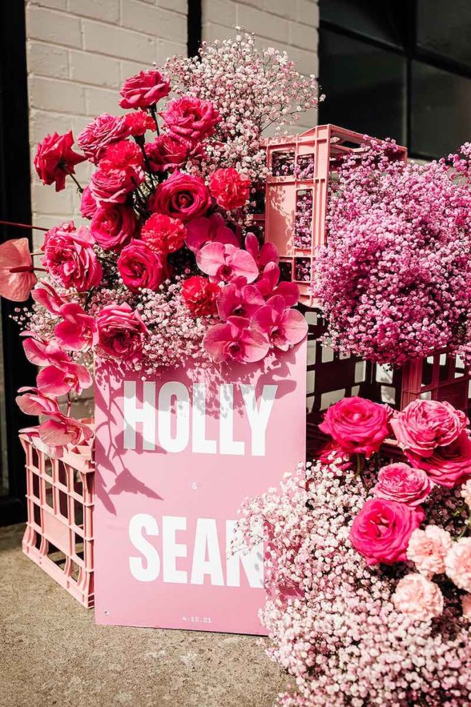 In questa foto un welcome table mariage per un matrimonio stile Barbie con un cartello di colore rosa con i nomi degli sposi Holly e Sean con rose, orchidee e gypsophila di colore rosa e fucsia