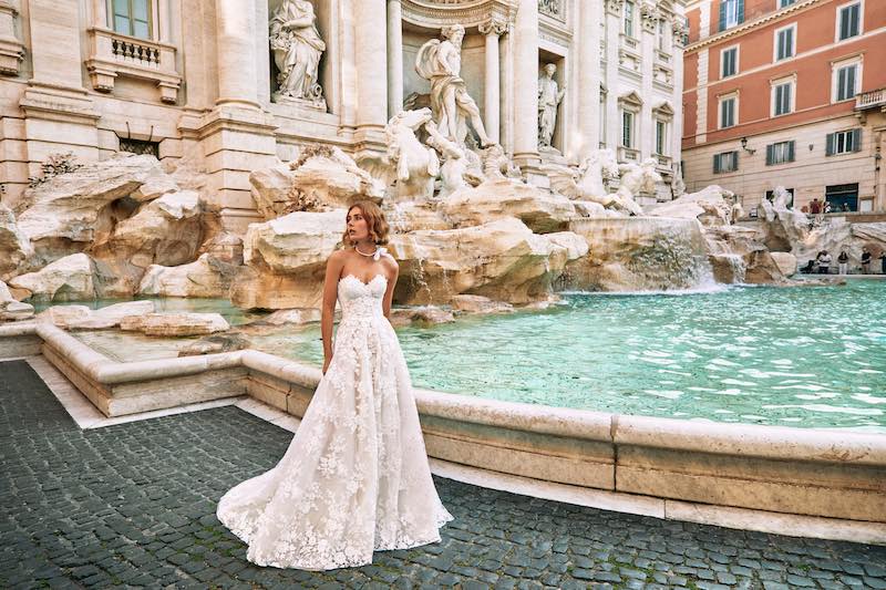 In questa foto una modella posa davanti alla Fontana di Trevi indossando uno degli abiti da sposa romantici 2024 di Atelier Emé: un modello con scollo a cuore, gonna a 3  e pizzo con ricami floreali 3D all over