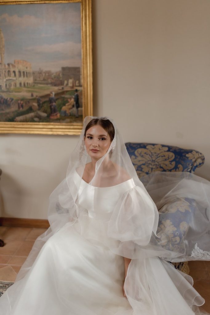 In questa foto una sposa seduta su una poltrona d'epoca a Villa del Gattopardo indossa un abito in satin e un velo di tulle 