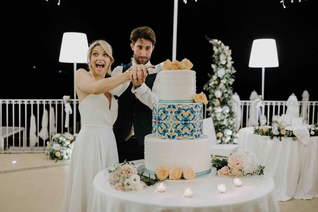 In questa immagine gli sposi tagliano la torta a tre piani, con al centro un piano con il decoro tipico delle maioliche siciliane.