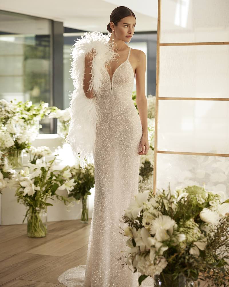 In questa foto una modella posa indossa un abito da sposa Rosa Clará Couture 2024 modello lingerie ricamato con perline all over, con scollo a V, spalline sottili, abbinato ad un coprispalle di piume